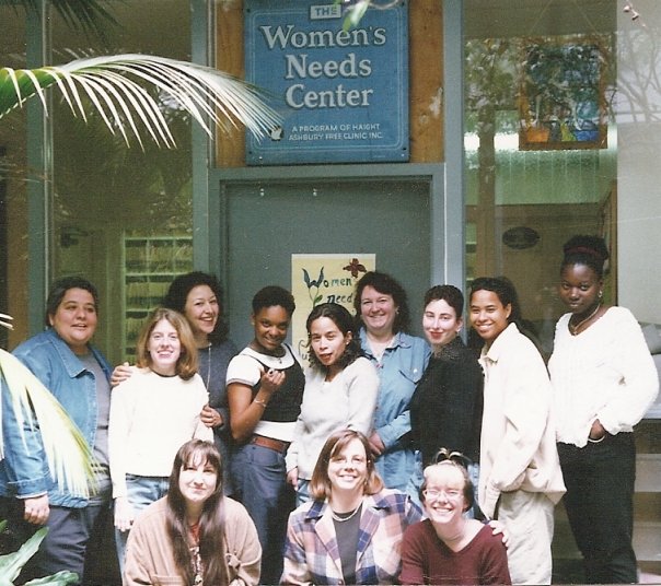 Group color photo of a diverse group of women in casual wear in front of a door with a sign that reads, "Women's Needs Center.'