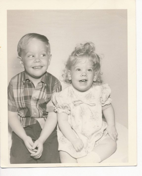 B/w photo of a little boy and younger girl sitting. Both smile and the boy is giving the girl a mischievous sideways glance.