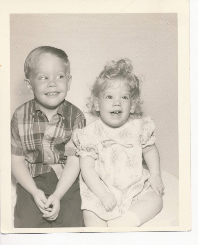 B/w photo of a little boy and younger girl sitting. Both smile and the boy is giving the girl a mischievous sideways glance.