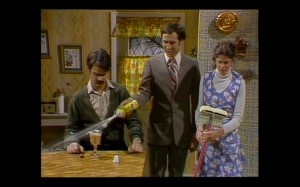 Color still from Saturday Night Live. A parody commercial for Shimmer Floor Wax. Chevy Chase is spraying Shimmer on Dan Ackroyd's pudding while Gilda Radner looks at the Shimmer on her mop.