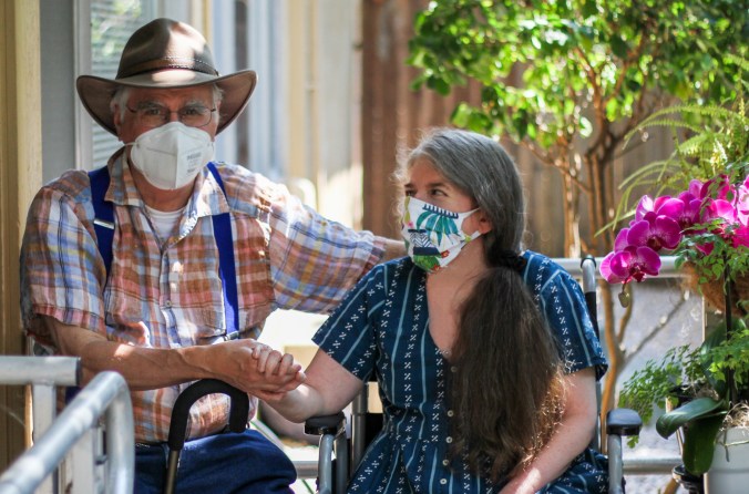 An older white man in a hat and face mask, holding the hand of a middle-aged white woman in a wheelchair, as she is turned to smile at him